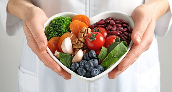 a woman holding a heart shaped bowl of fruit, vegetables, nuts, beans and greens